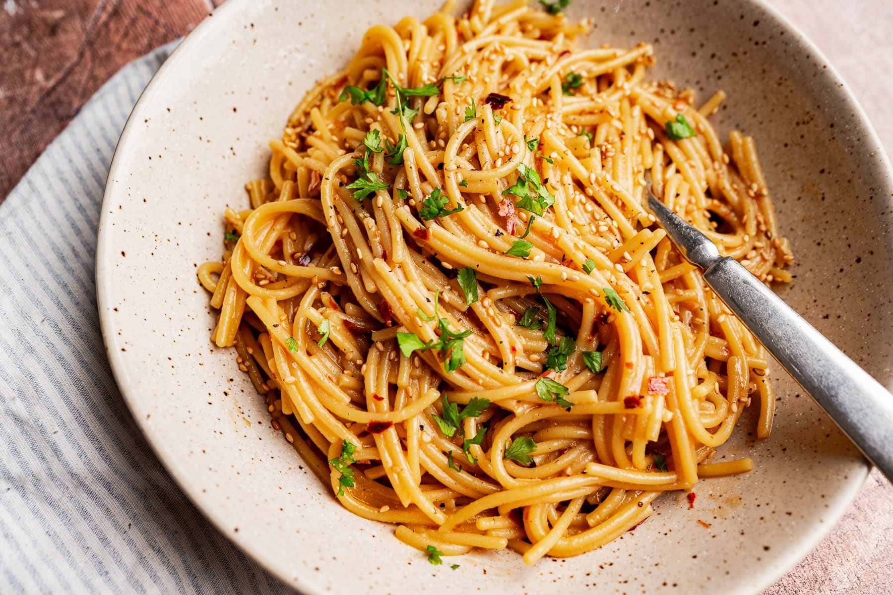A bowl of chili oil pasta with cooked spaghetti noodles, topped with chopped herbs and sesame seeds, with a fork placed inside the bowl.