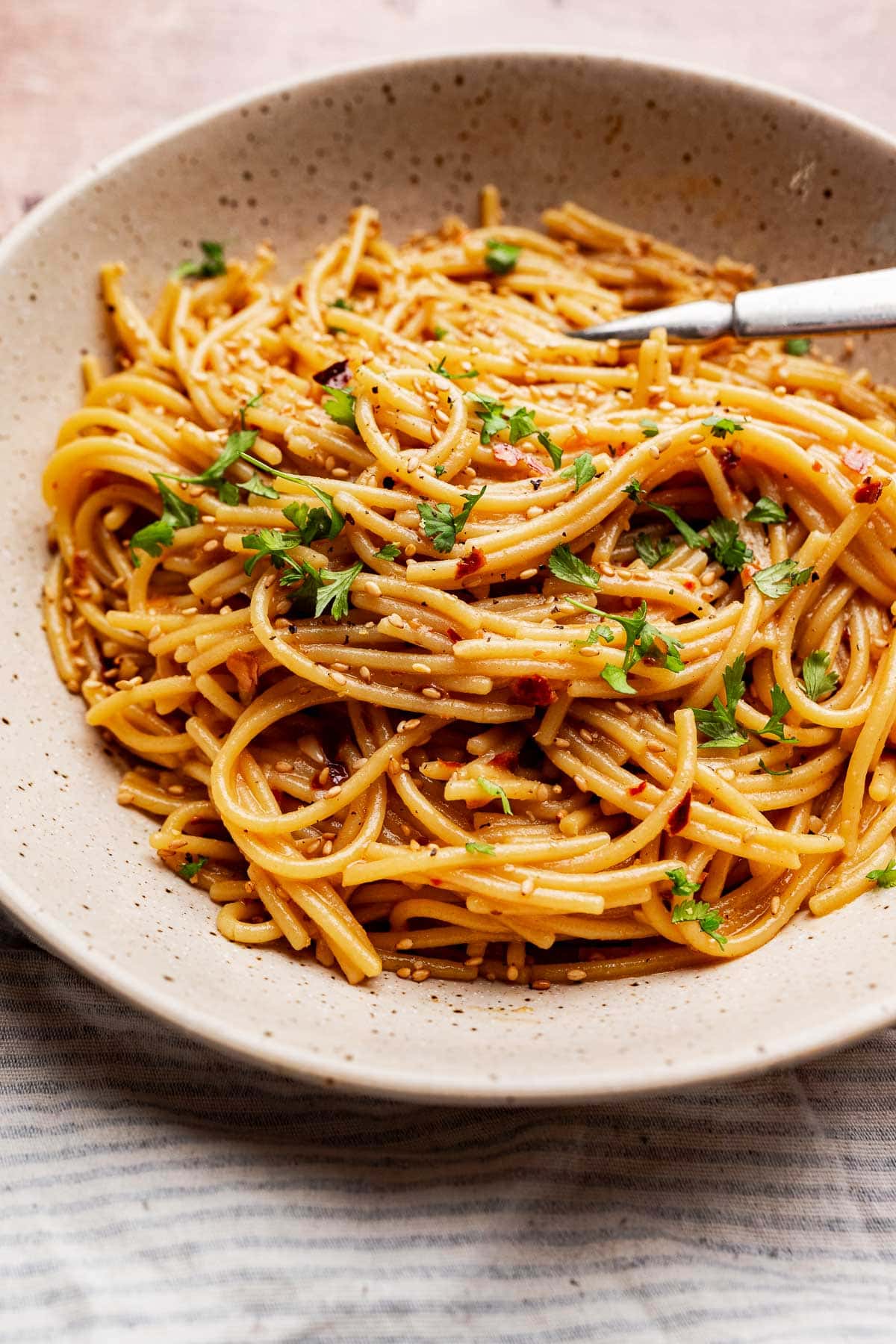 A beige ceramic bowl filled with chili oil pasta—spaghetti noodles topped with chopped parsley and sesame seeds, with a fork resting on the edge.