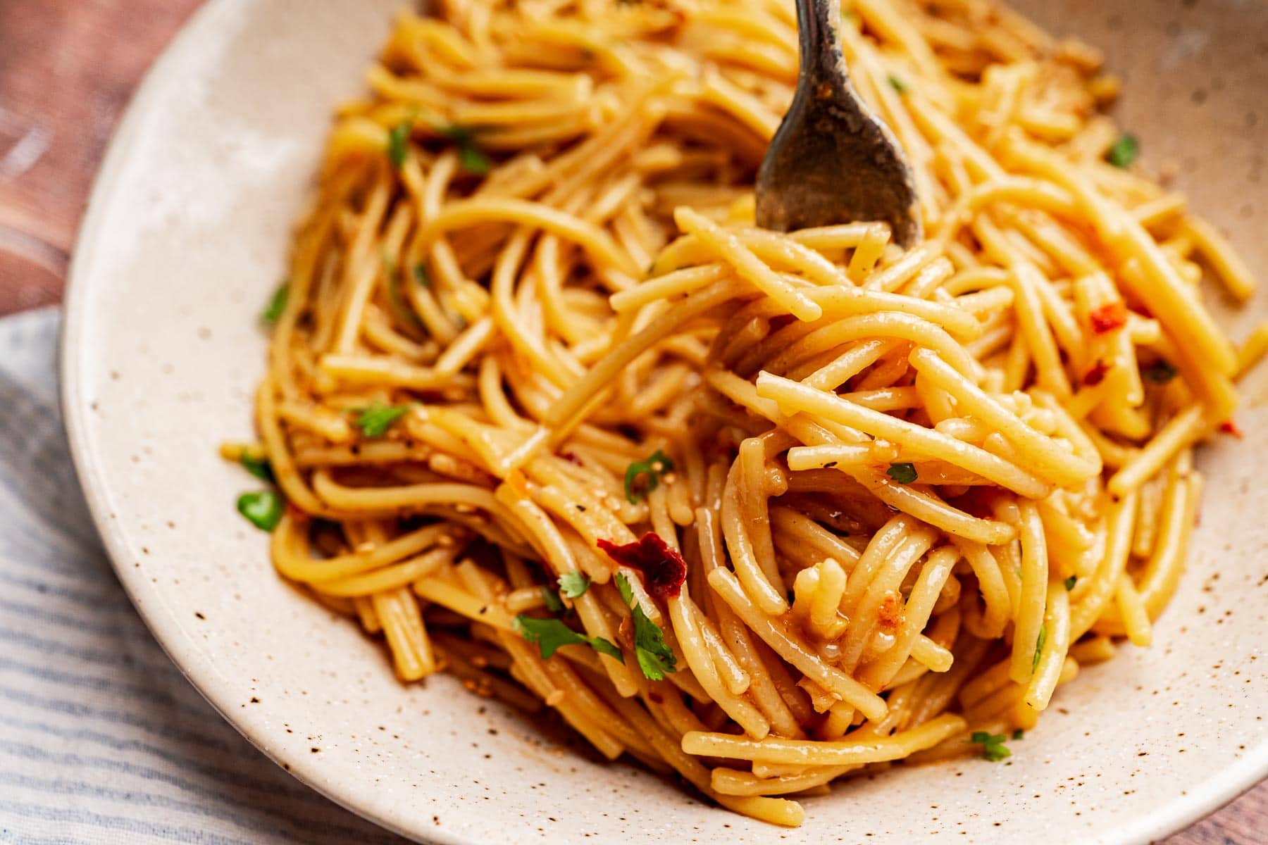 Close-up of a plate of cooked chili oil pasta with a fork twirling noodles, garnished with herbs and a few pieces of sundried tomato.