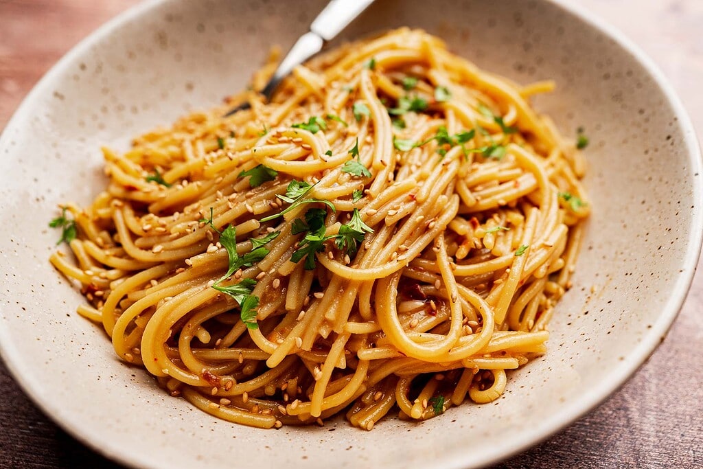 A bowl of cooked noodles tossed in chili oil pasta sauce, garnished with chopped herbs and sesame seeds, with a fork resting on the side.