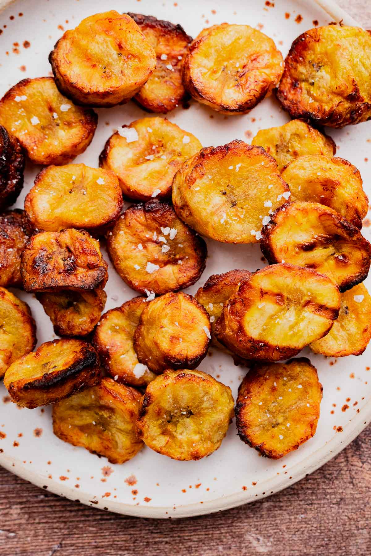 A plate of sliced, golden-brown air fryer sweet plantains sprinkled with coarse salt rests on a white plate.
