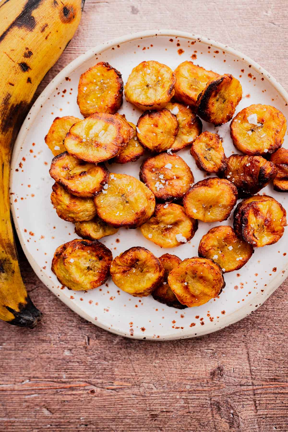 A plate of sliced, roasted air fryer sweet plantains sprinkled with salt, with a whole plantain beside the plate on a wooden surface.