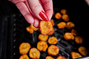 A hand with red nail polish holds up an air fryer sweet plantain chip above a tray of similar chips.