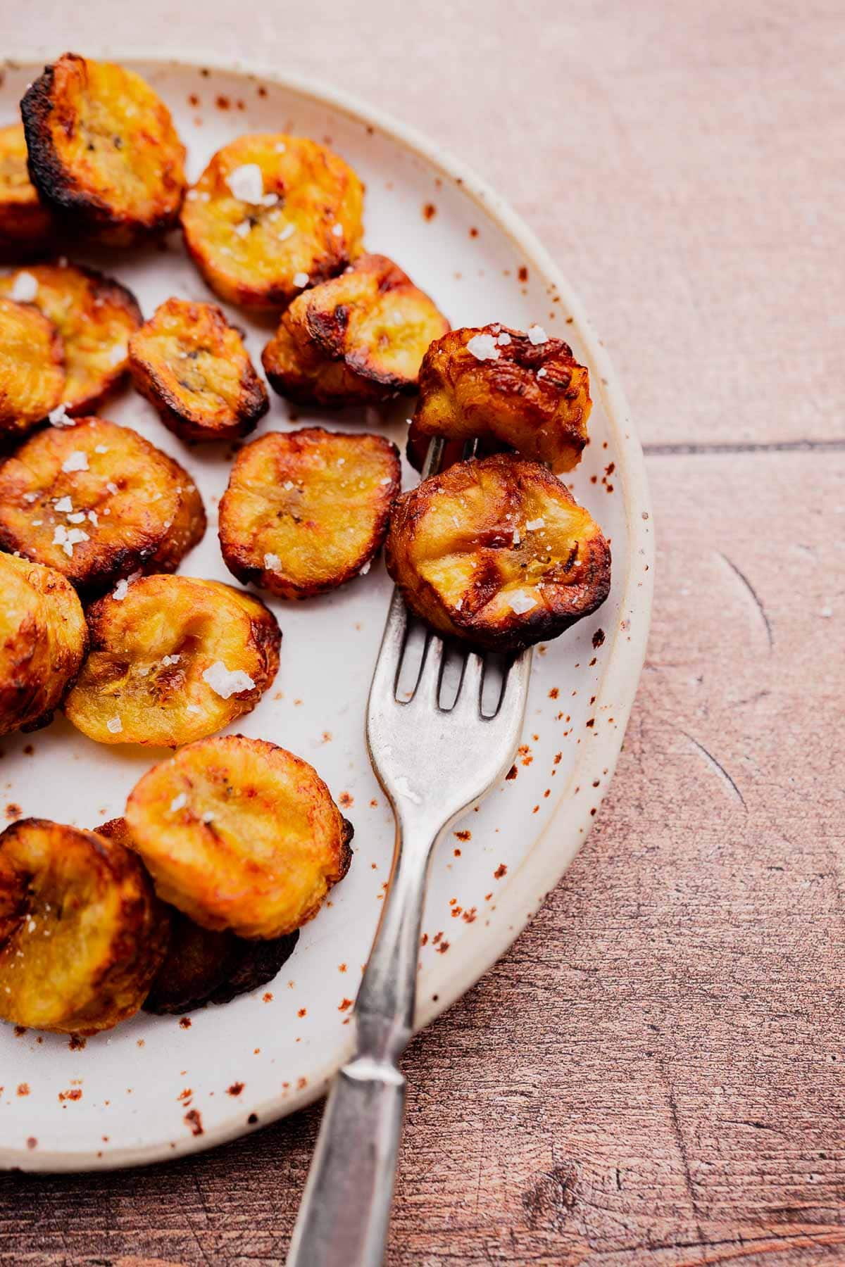 A plate of air fryer sweet plantains, sliced and cooked to perfection with a sprinkling of coarse salt, accompanied by a fork on a wooden surface.