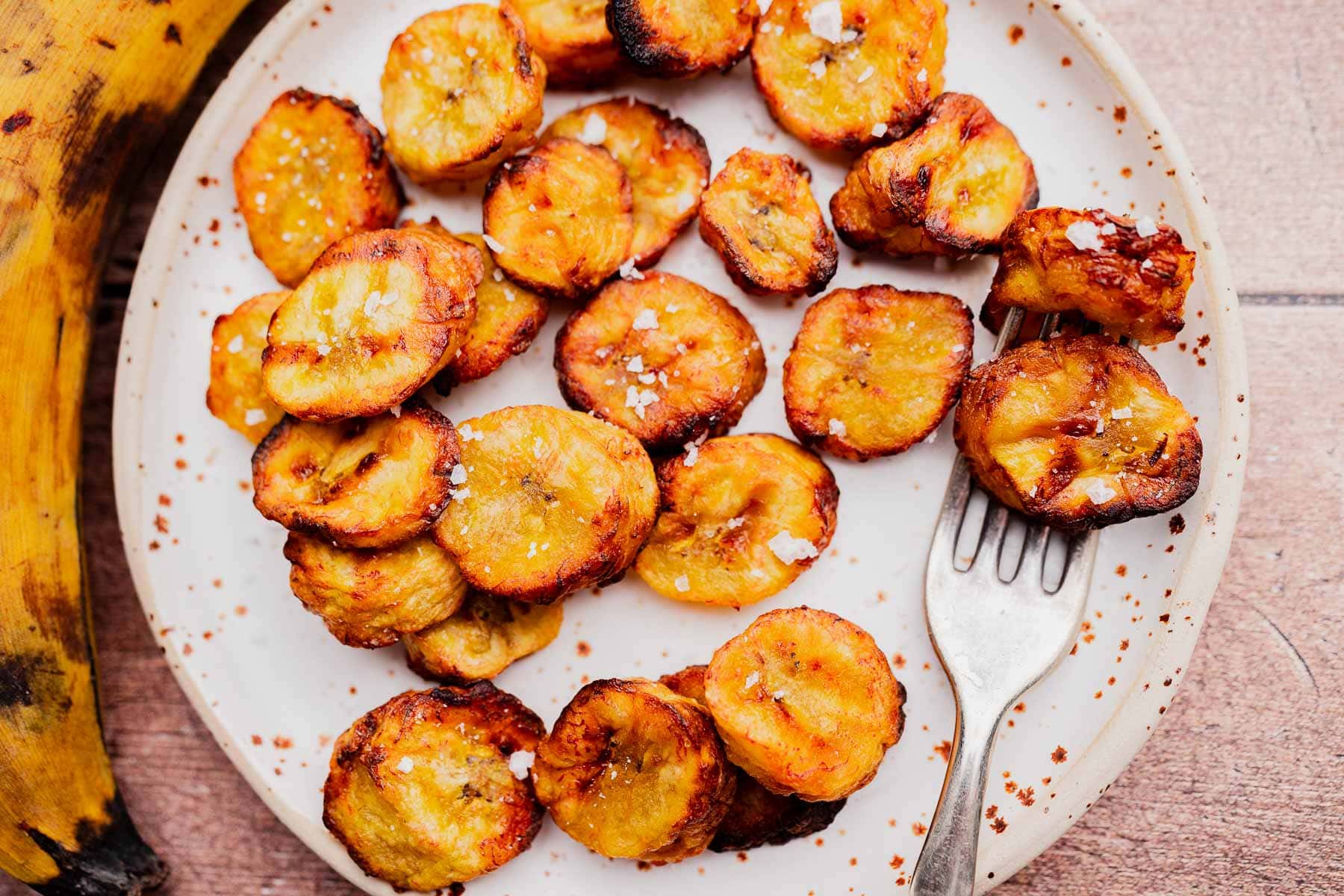 A plate of crispy, golden-brown air fryer sweet plantains topped with coarse salt, with a fork resting on the plate.