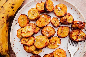 A plate of crispy air fryer sweet plantains slices sprinkled with coarse salt, with a fork on the side and a whole plantain partially visible.