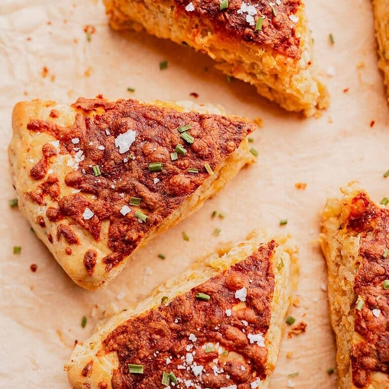 Three triangular gluten free scones topped with melted cheese, coarse salt, and chopped chives on parchment paper.