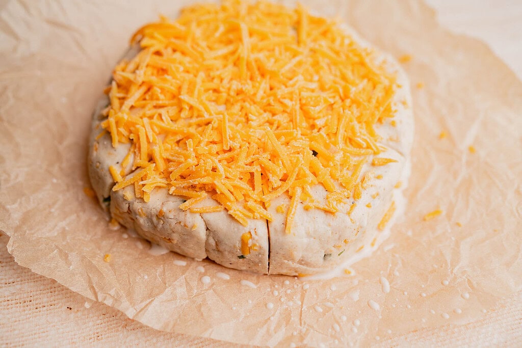 Unbaked round gluten free scones topped with shredded cheddar cheese sit on parchment paper, ready for baking.