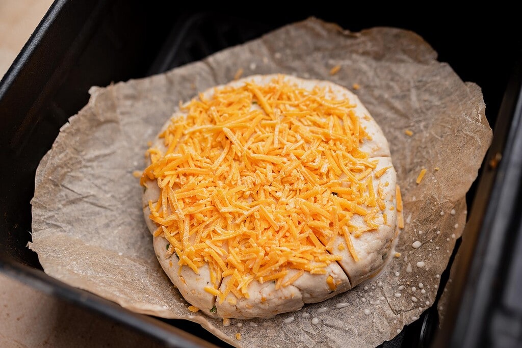 A round, unbaked gluten free scone dough topped with shredded cheddar cheese sits on parchment paper inside a baking tray.
