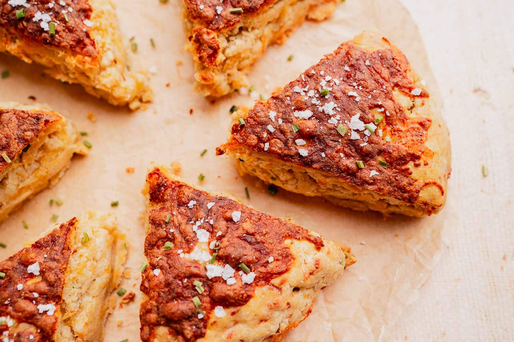 Triangular pieces of gluten free scones topped with herbs and flaked salt are arranged on parchment paper.