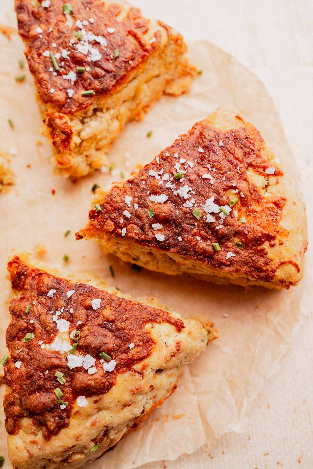 Three triangular gluten free scones, topped with flaky sea salt and chopped herbs, are arranged on a piece of parchment paper.