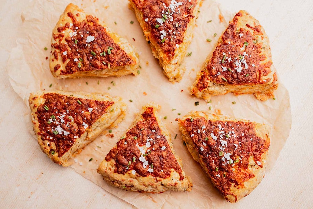 Six triangular, golden-brown gluten free scones with a cheesy topping are arranged in a circle on parchment paper, sprinkled with herbs and coarse salt.