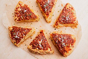Six triangular, golden-brown gluten free scones with a cheesy topping are arranged in a circle on parchment paper, sprinkled with herbs and coarse salt.