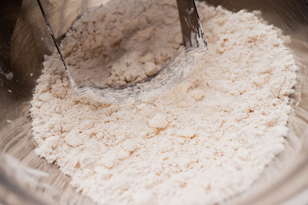A pastry cutter blends flour and fat in a metal bowl, creating a coarse, crumbly mixture—ideal for making gluten free scones.