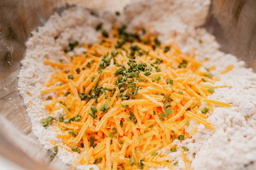 Shredded cheddar cheese and chopped chives on top of a flour mixture in a metal mixing bowl, perfect for making gluten free scones.