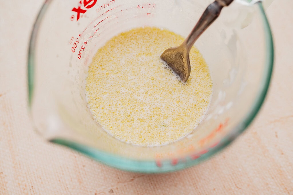 A glass measuring cup filled with a yellow liquid mixture for gluten free scones being stirred by a metal spoon.