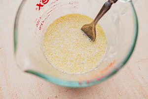 A glass measuring cup filled with a yellow liquid mixture for gluten free scones being stirred by a metal spoon.