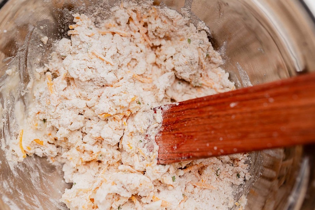 A wooden spatula stirs a mixture of flour, shredded cheese, and other baking ingredients in a metal bowl for gluten free scones.