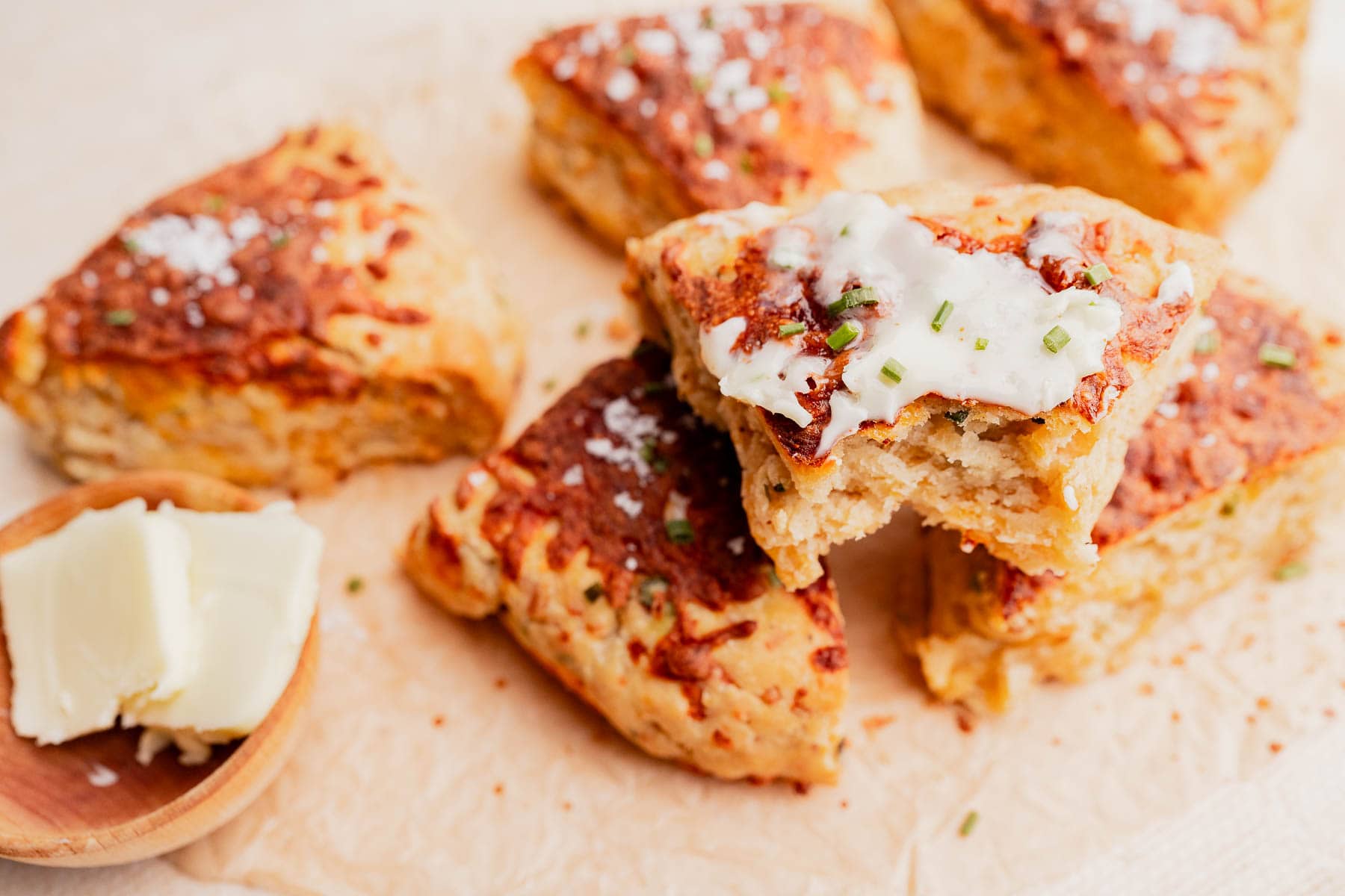 Gluten free scones topped with butter and chives rest on parchment paper, while a wooden dish holds two butter pats nearby.