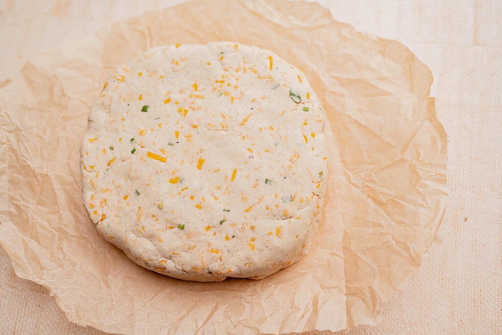 A round, uncooked gluten free scone dough with visible cheese and herb pieces sits on a sheet of crumpled parchment paper.