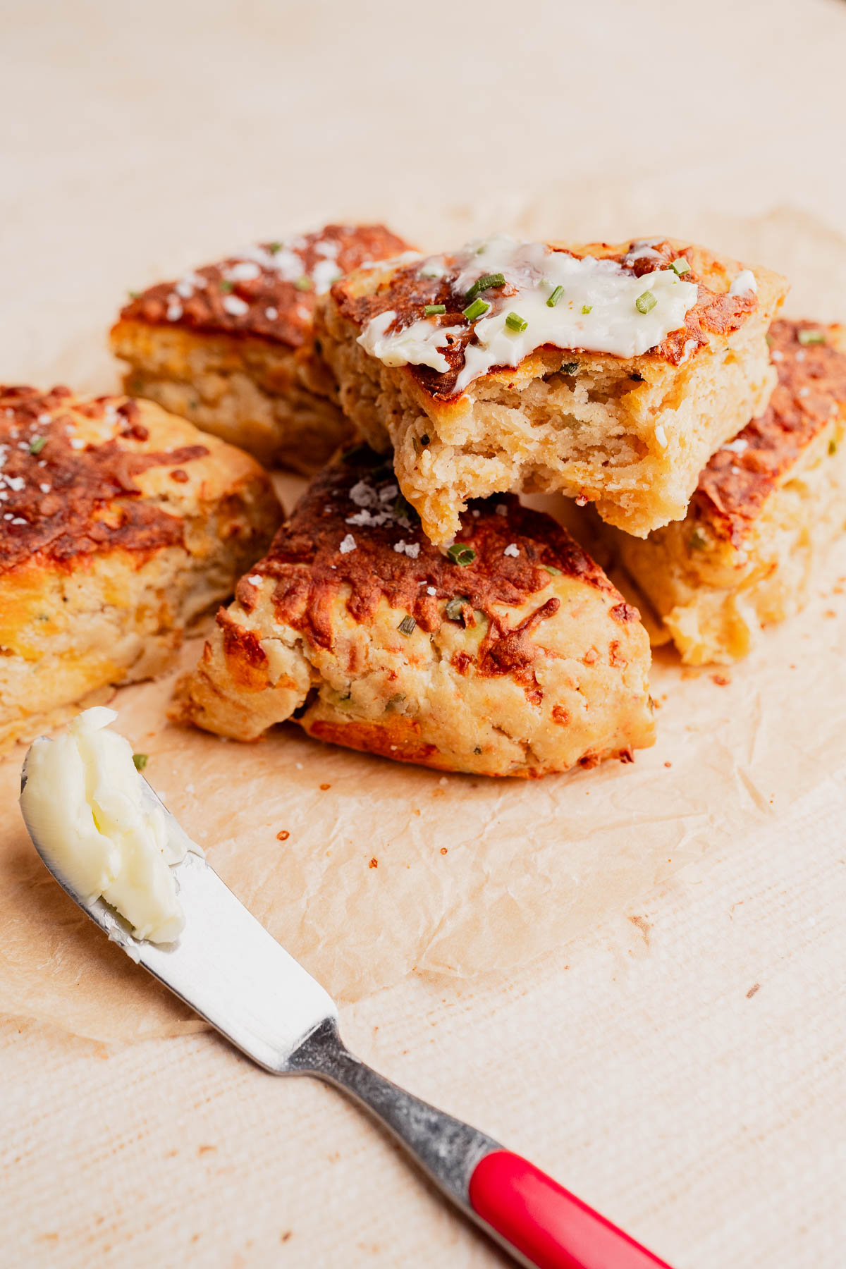 Gluten free scones topped with butter and herbs are stacked on parchment paper, with a butter knife resting nearby.
