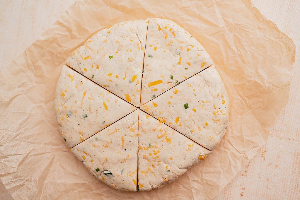Round disc of unbaked gluten free scones dough with cheese and herbs, cut into six wedges, resting on a sheet of parchment paper.