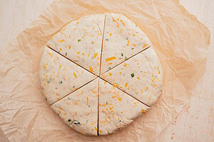 Round disc of unbaked gluten free scones dough with cheese and herbs, cut into six wedges, resting on a sheet of parchment paper.