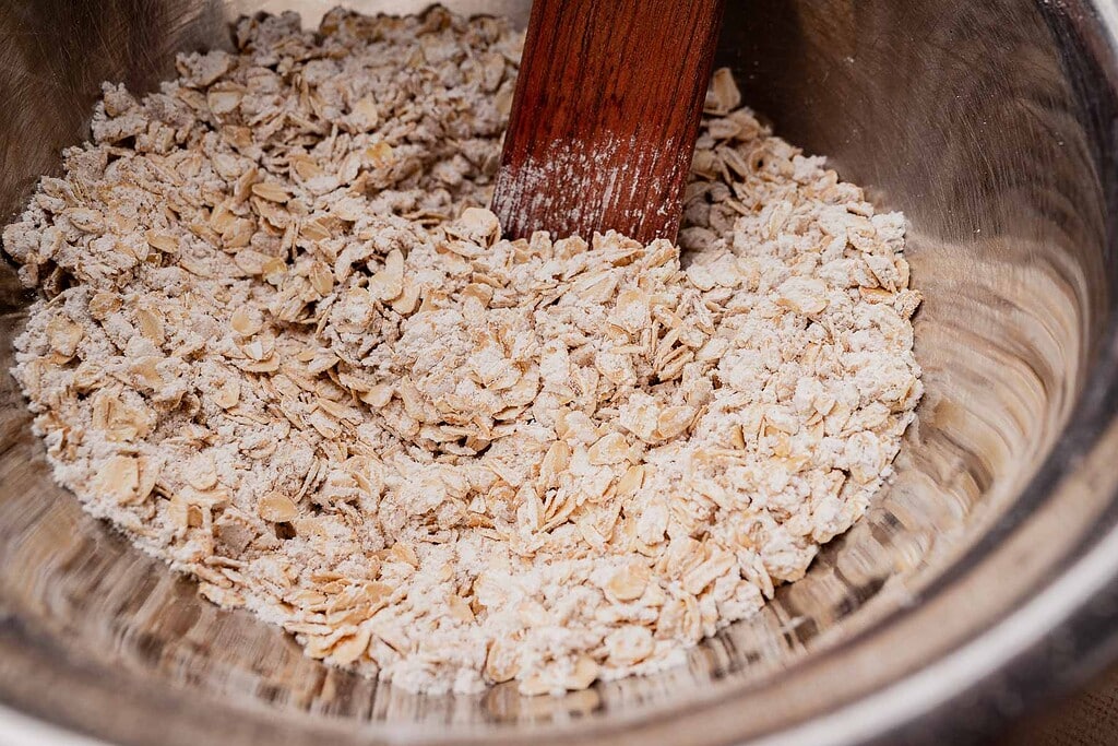 A wooden spatula mixes rolled oats and flour in a stainless steel bowl, prepping the base for gluten free oatmeal chocolate chip cookies.