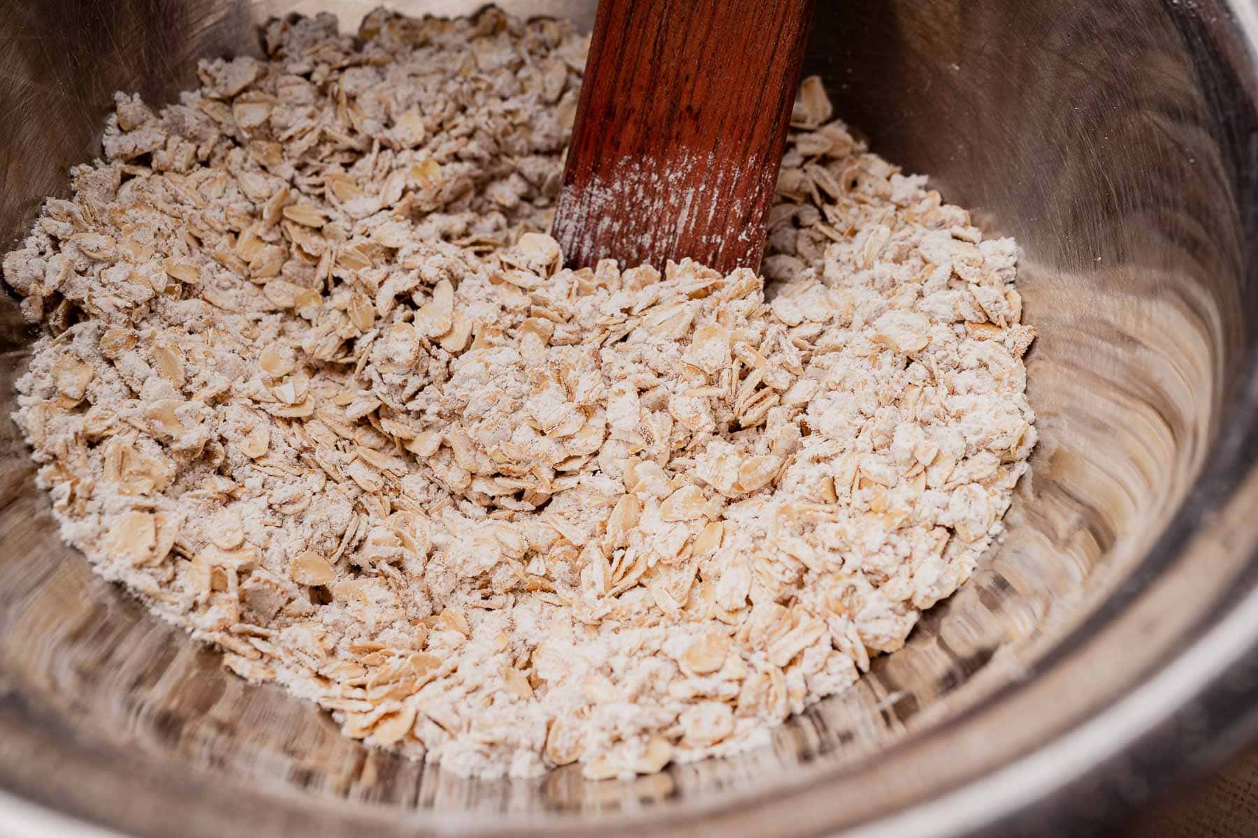 A close-up of rolled oats mixed with flour in a metal bowl, being stirred with a wooden spatula while preparing gluten free oatmeal chocolate chip cookies.