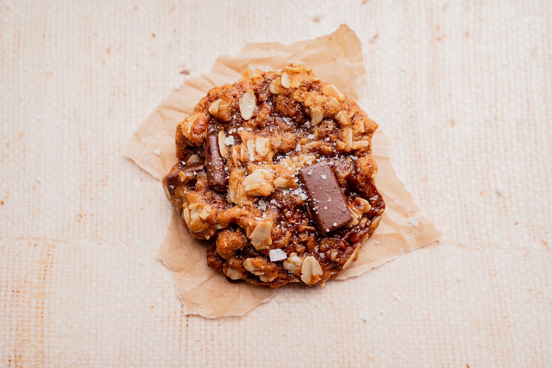 A single gluten free oatmeal chocolate chip cookie on a piece of brown parchment paper, photographed from above on a light textured surface.