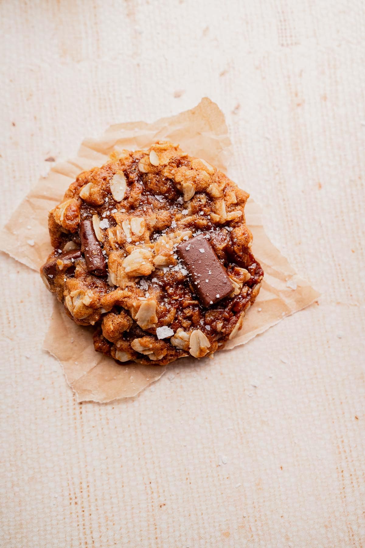 A gluten free oatmeal chocolate chip cookie with visible oats and chocolate pieces rests on a crumpled piece of parchment paper atop a light textured surface.