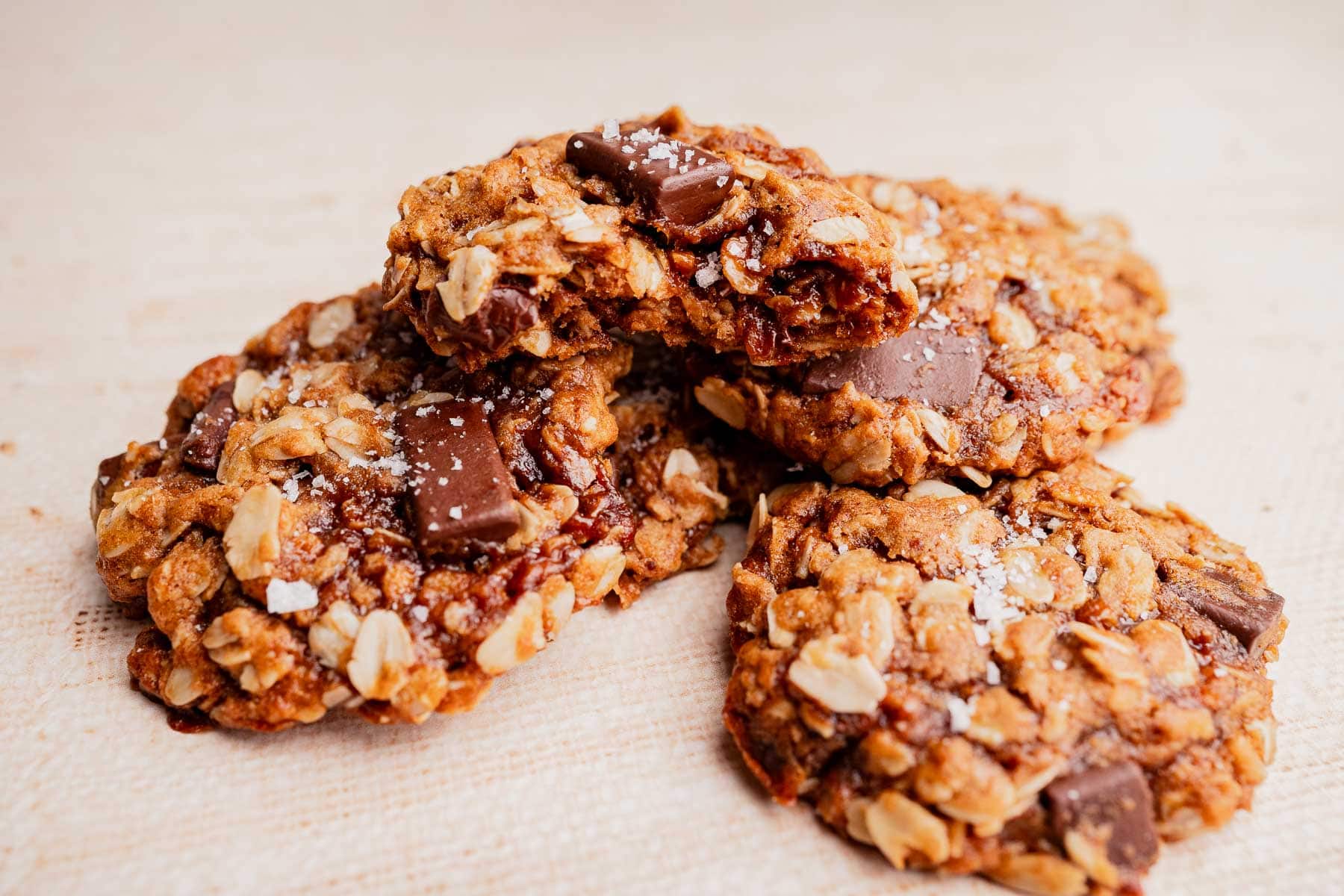 Three gluten free oatmeal chocolate chip cookies with visible chocolate chunks and sprinkled sea salt rest on a light surface.