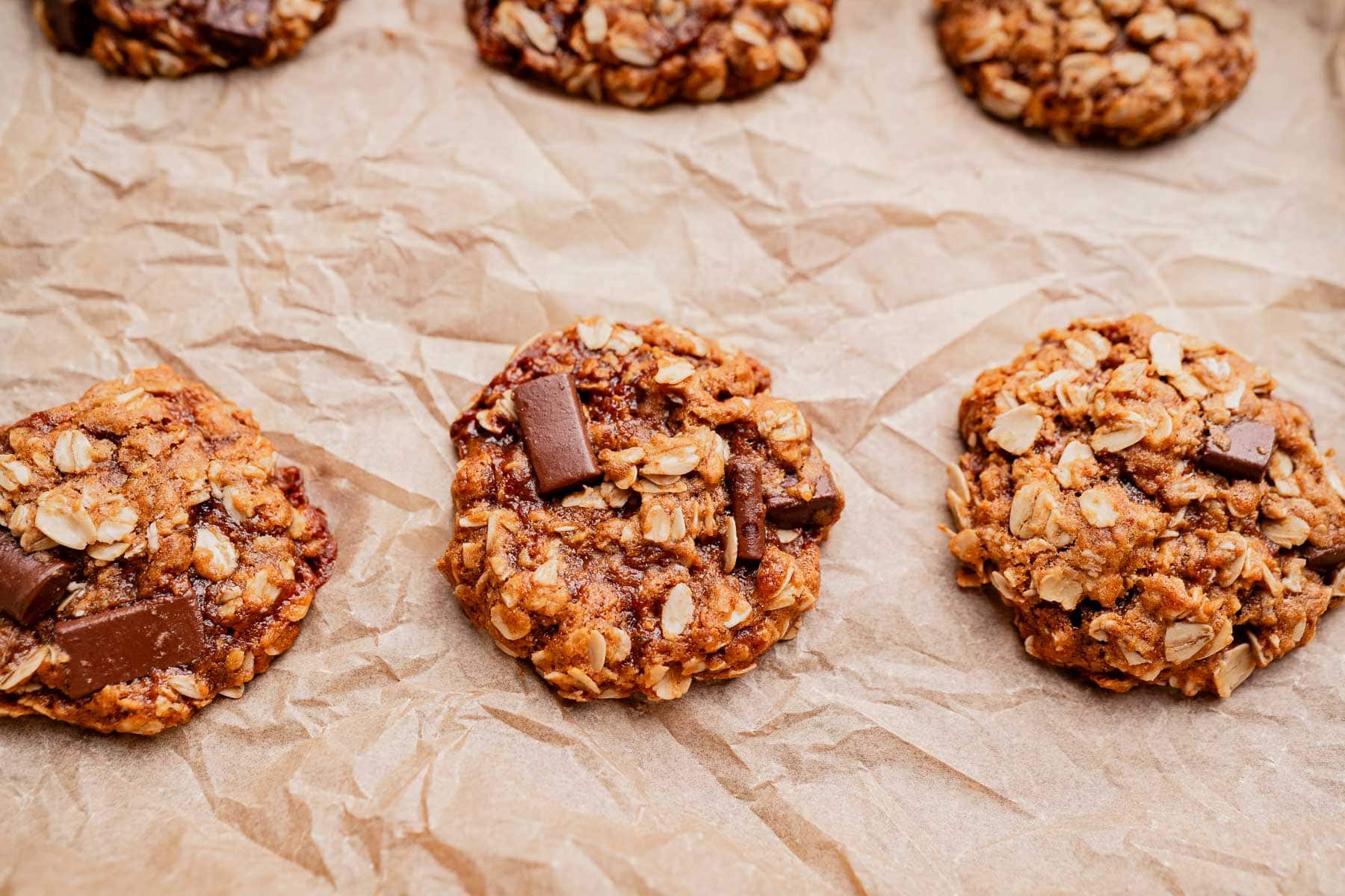 Three gluten free oatmeal chocolate chip cookies rest on crumpled parchment paper, with more cookies partially visible in the background.