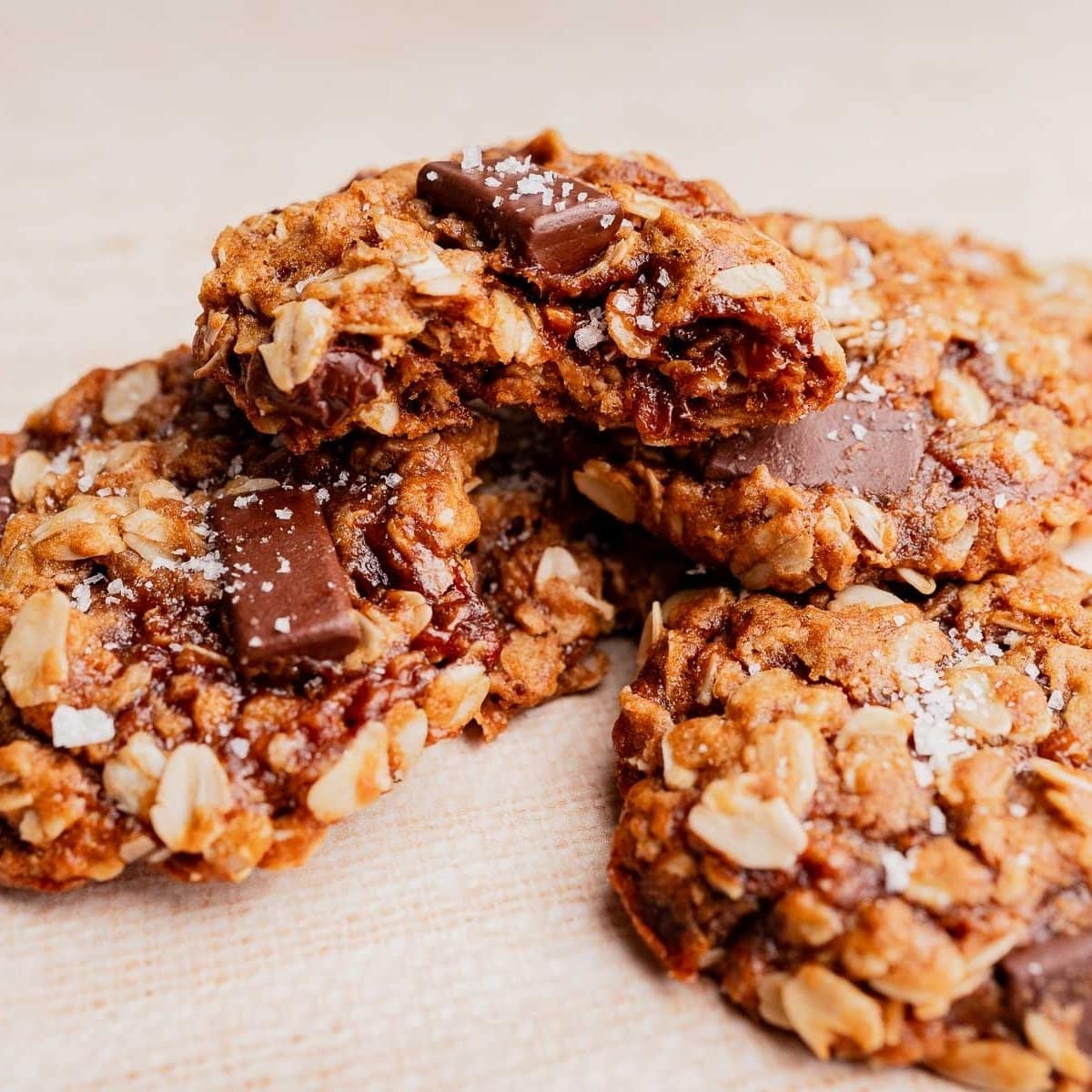 Three gluten free oatmeal chocolate chip cookies with visible oats and chocolate chunks, sprinkled with sea salt, stacked on a light surface.
