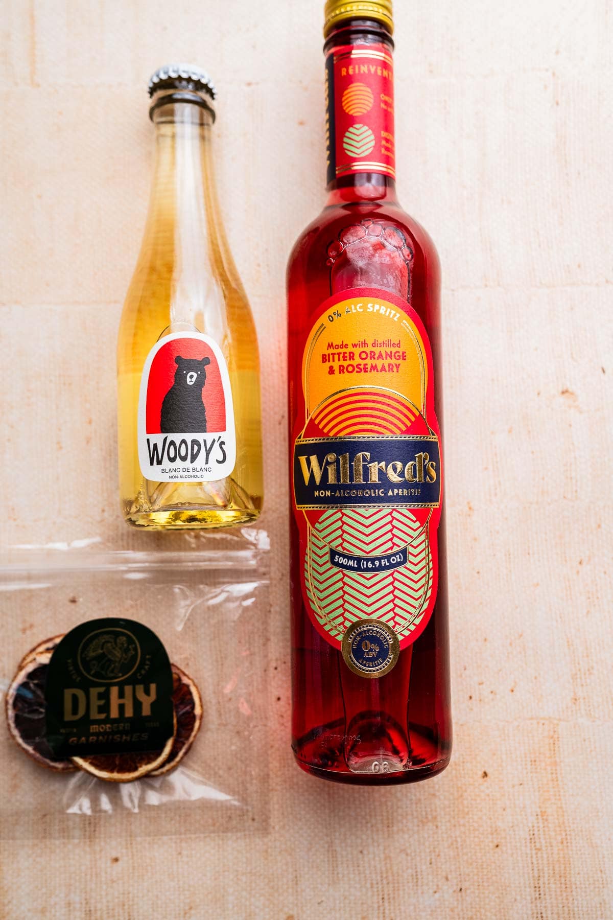 A bottle of Woody’s apple juice, a bottle of Wilfred’s bitter orange & rosemary aperitif—perfect for crafting an aperol spritz mocktail—and a packet of dried orange slices on a light surface.