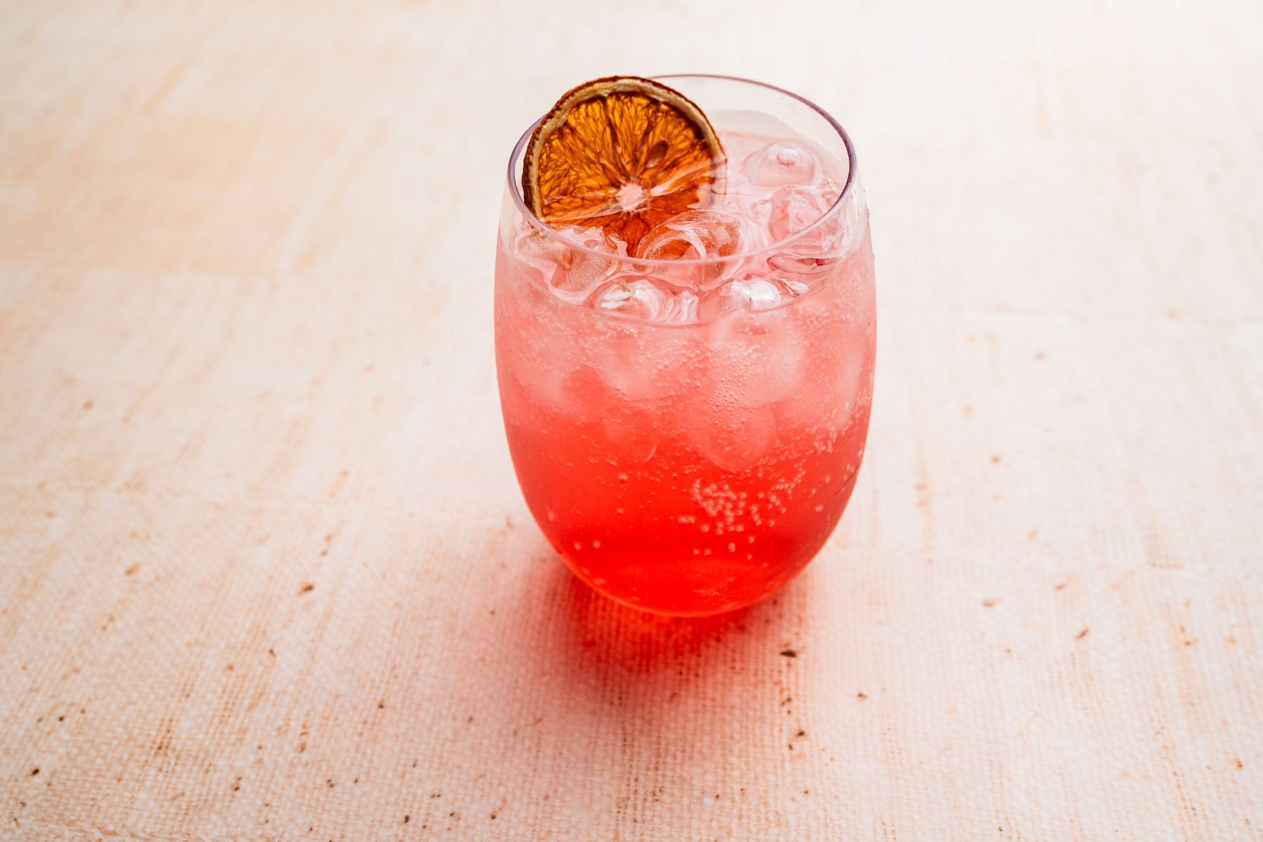 A glass of red aperol spritz mocktail, topped with a slice of dried citrus, sits elegantly on a light-colored surface.