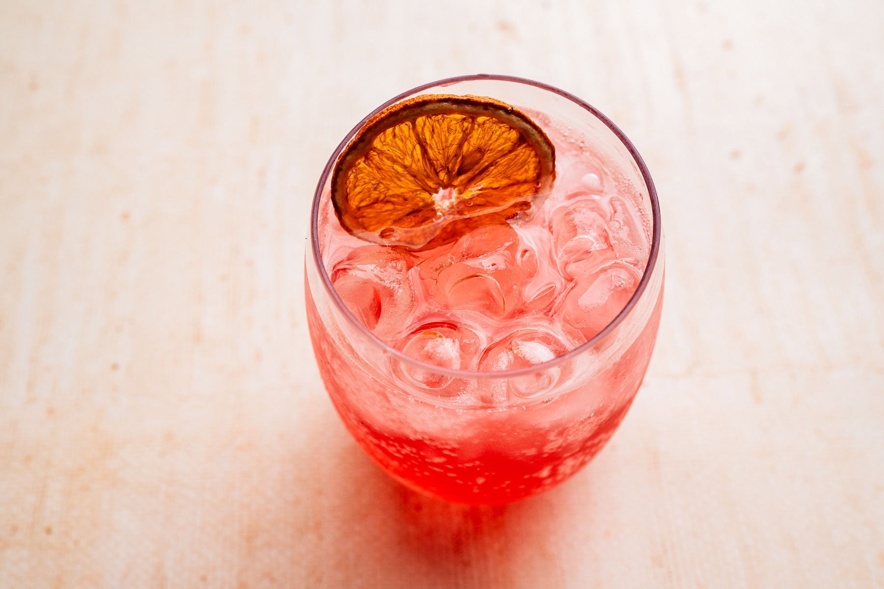 A round glass filled with a pink aperol spritz mocktail, topped with a dried orange slice, sits on a light wooden surface.