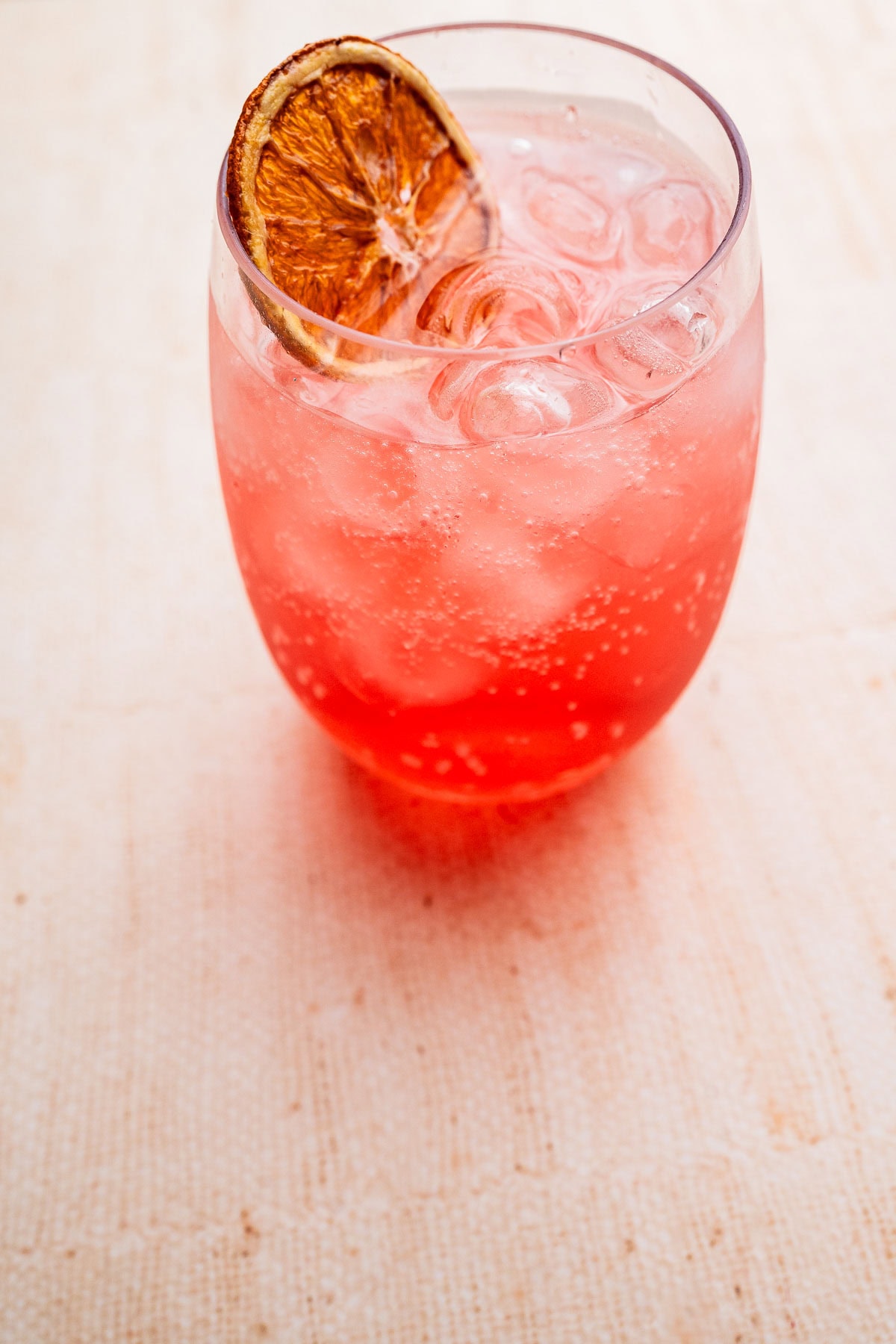 A glass of pink sparkling aperol spritz mocktail with ice cubes and a dried orange slice garnish on a light-colored surface.