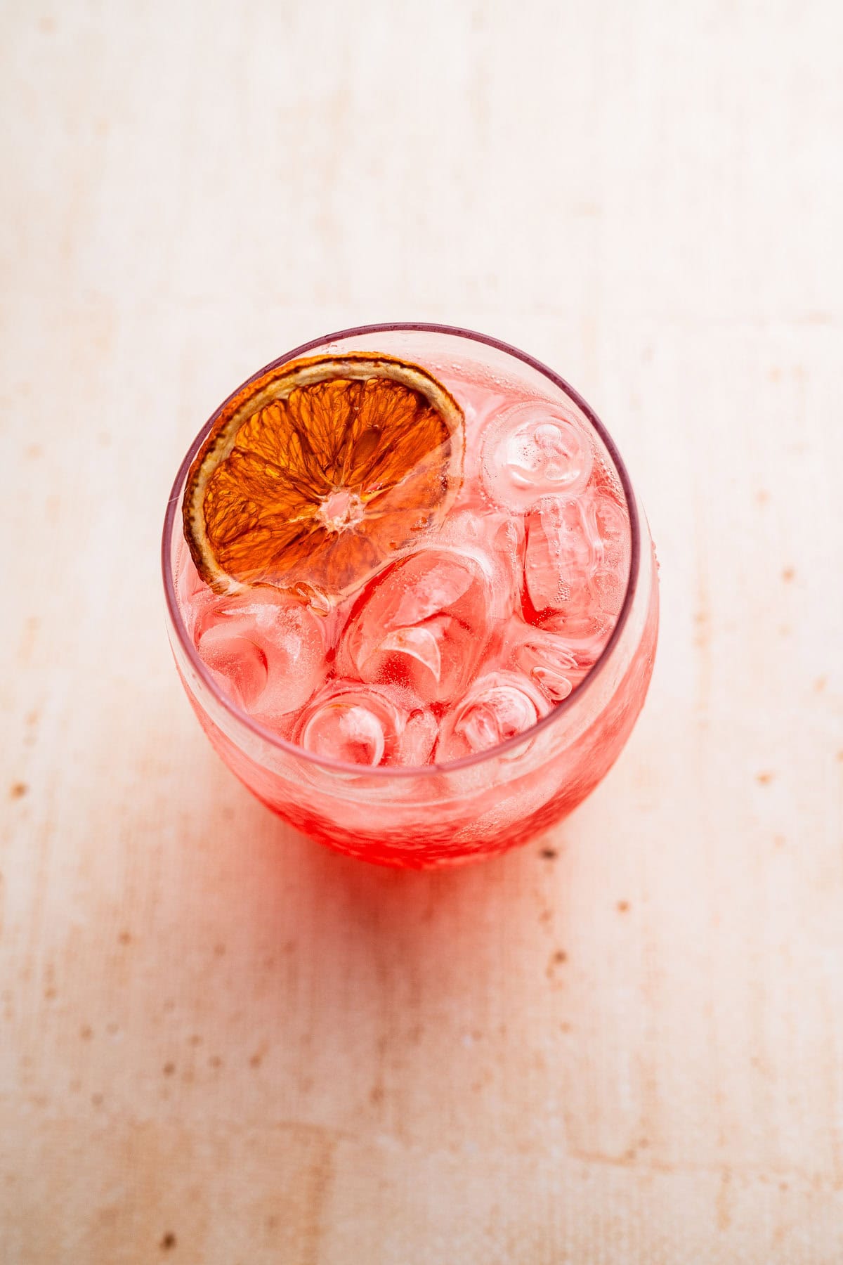 A round glass filled with a pink Aperol Spritz mocktail, garnished with a dried citrus slice, sits on a light-colored surface.