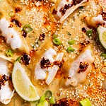 Close-up of baked dumplings in a creamy sauce, topped with chili oil, sesame seeds, sliced green onions, and lime wedges.