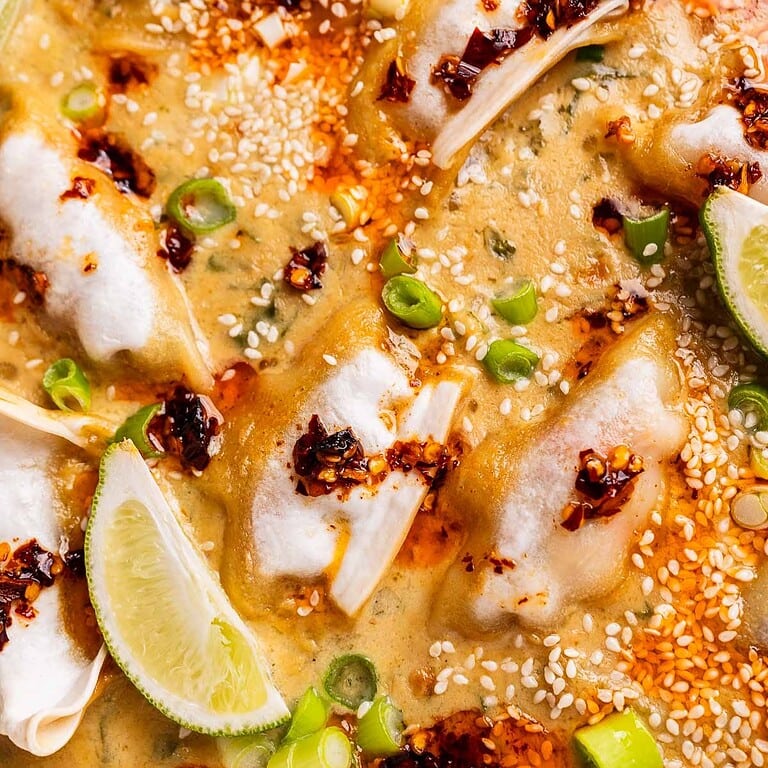 Close-up of baked dumplings in a creamy sauce, topped with chili oil, sesame seeds, sliced green onions, and lime wedges.
