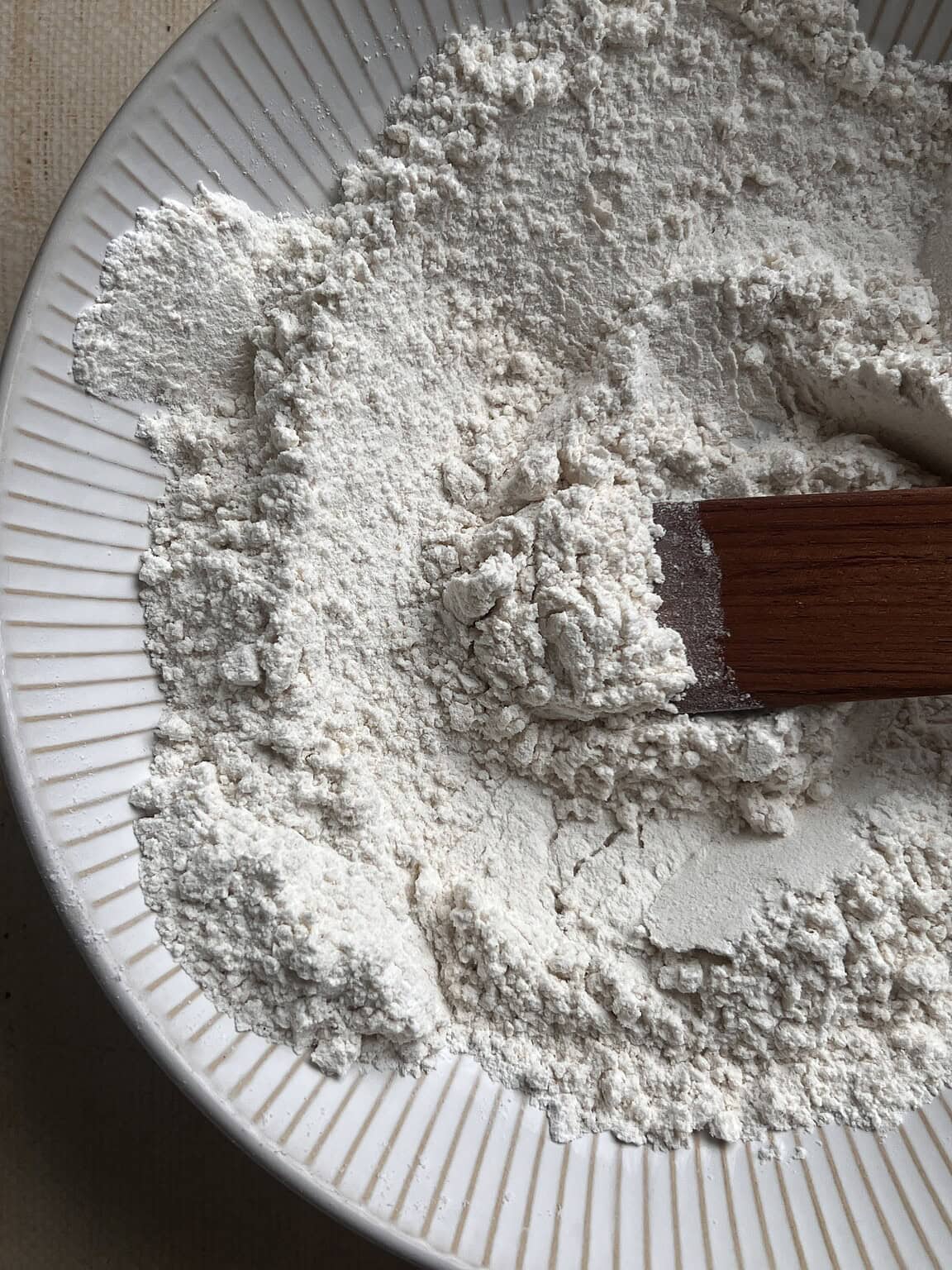 A close-up of white flour being mixed with a wooden spatula in a white, ridged bowl, perfect for making gluten free butter swim biscuits.