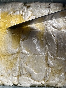 A baking dish filled with unbaked gluten free butter swim biscuits, divided into squares with a knife and topped with melted butter.