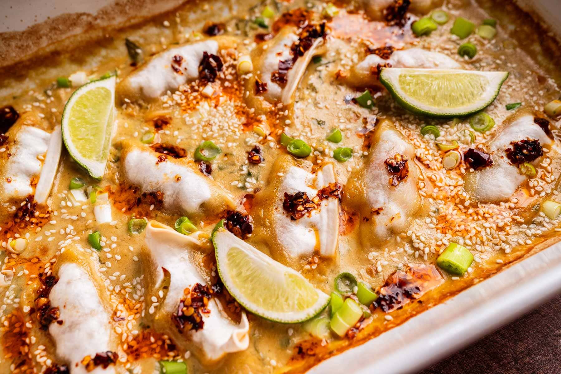 Baked dumplings in a creamy sauce, topped with chili oil, sesame seeds, chopped green onions, and fresh lime wedges in a rectangular dish. A delicious twist on classic baked dumplings bursting with flavor.