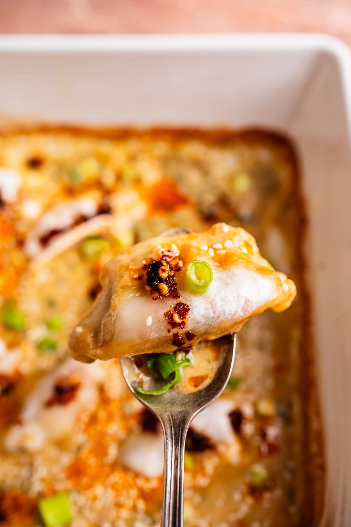 A close-up of baked dumplings topped with sauce and chopped green onions, held on a spoon above a casserole dish filled with more dumplings.
