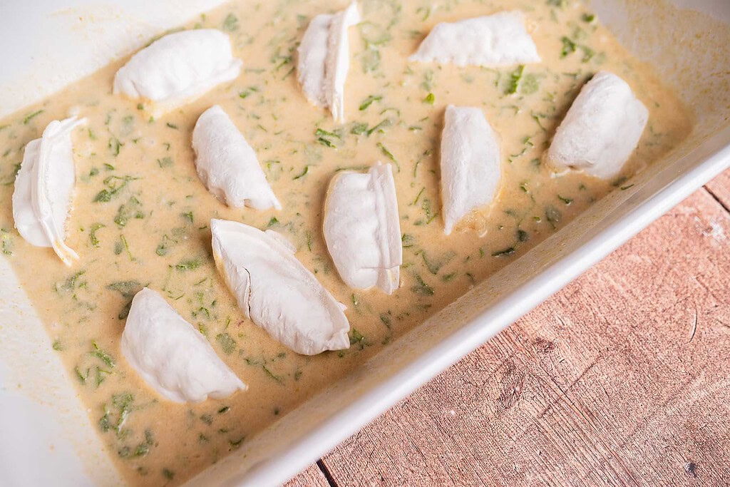 A white baking dish contains pieces of raw fish on top of a creamy, herb-filled sauce, alongside baked dumplings, placed on a wooden surface.