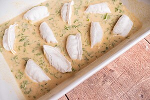 A white baking dish contains pieces of raw fish on top of a creamy, herb-filled sauce, alongside baked dumplings, placed on a wooden surface.