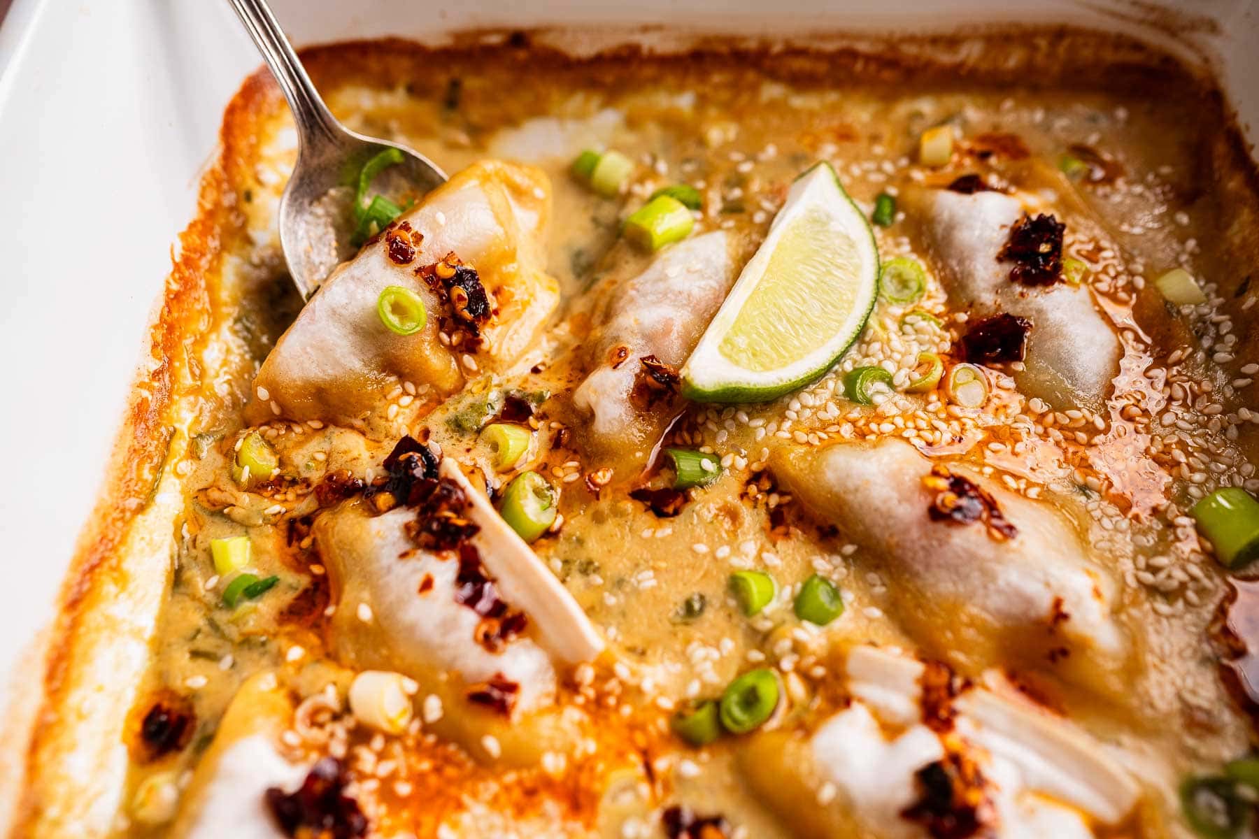 Close-up of baked dumplings in a creamy sauce, garnished with sliced green onions, sesame seeds, chili oil, and a lime wedge, with a spoon nestled among the baked dumplings.