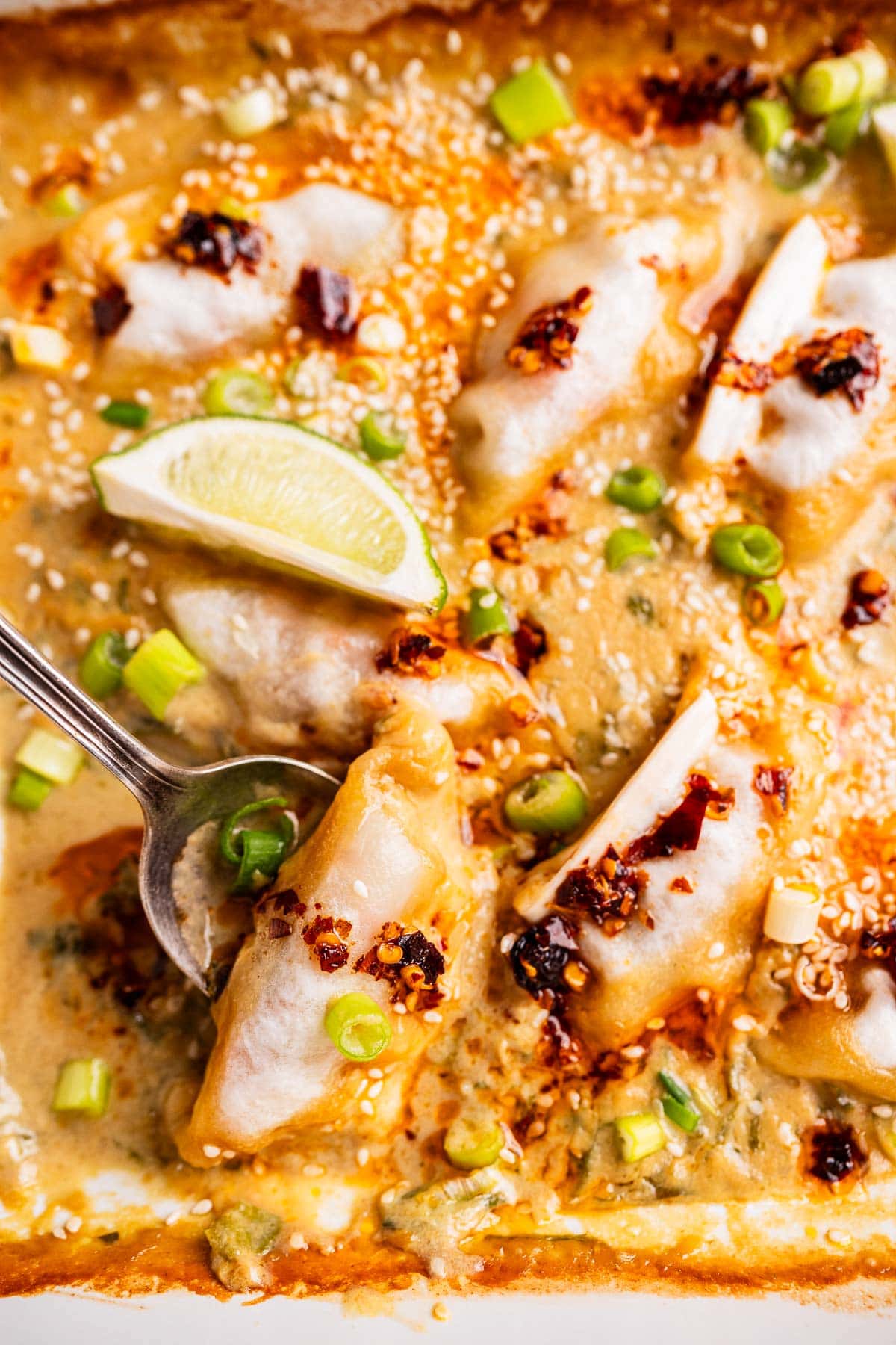 A close-up of baked dumplings topped with chili oil, sesame seeds, and chopped green onions, served with a wedge of lime; a spoon lifts one dumpling.