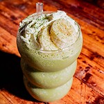 A matcha protein shake, topped with whipped cream, banana slices, and a sprinkle of green powder, served in a curvy glass on a wooden surface.